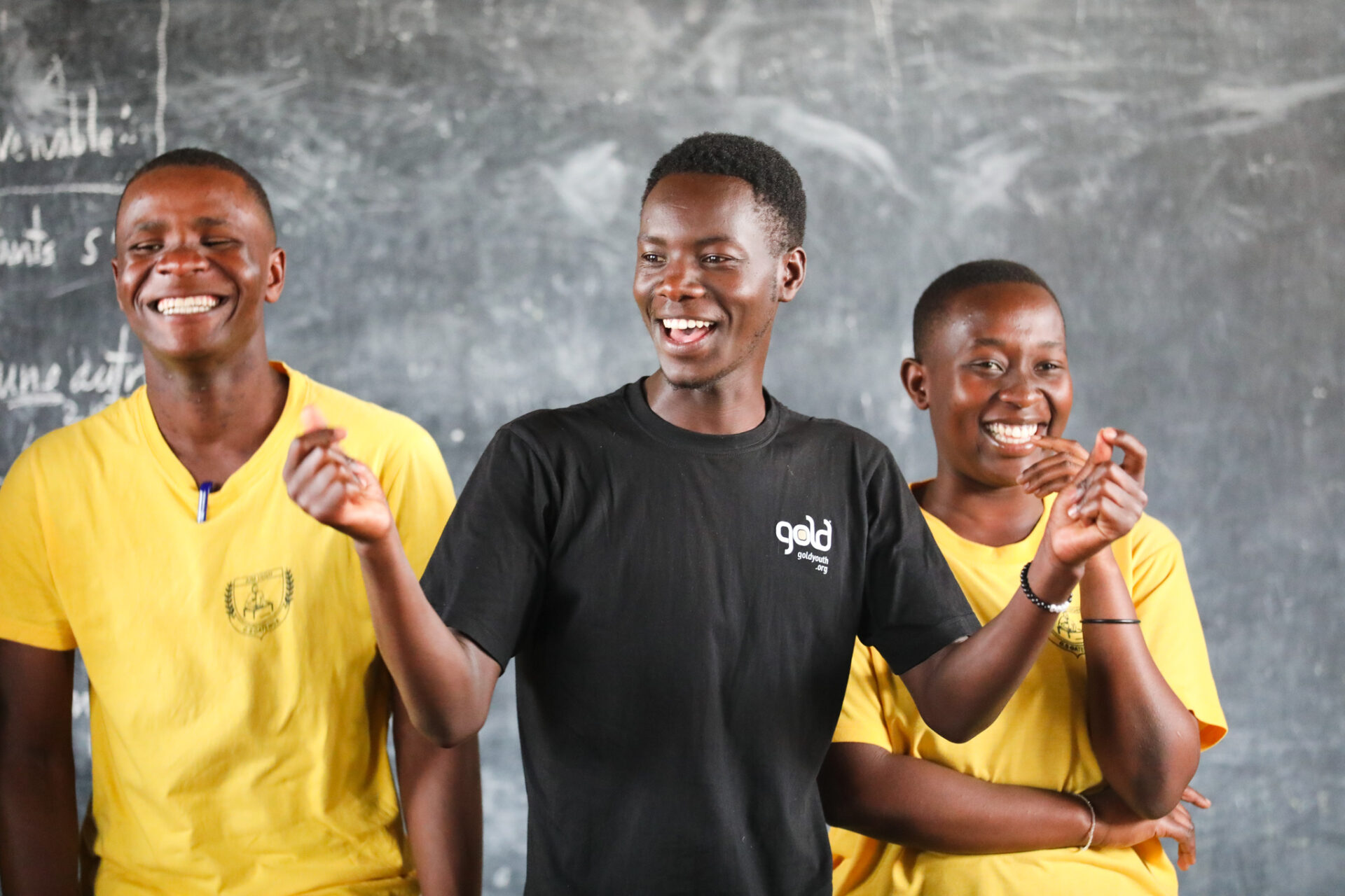 Three smiling gold-youth peer educators standing at a chalkboard during a session.